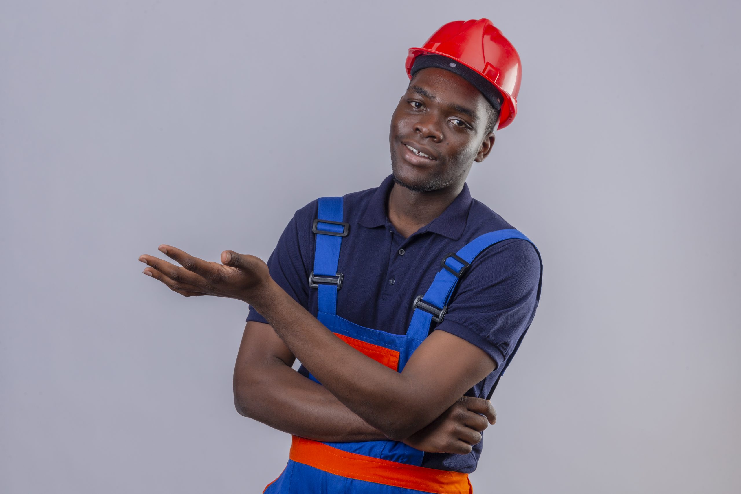 young african american builder man wearing construction uniform and safety helmet pointing with palm of hand looking at camera with smile standing over white background