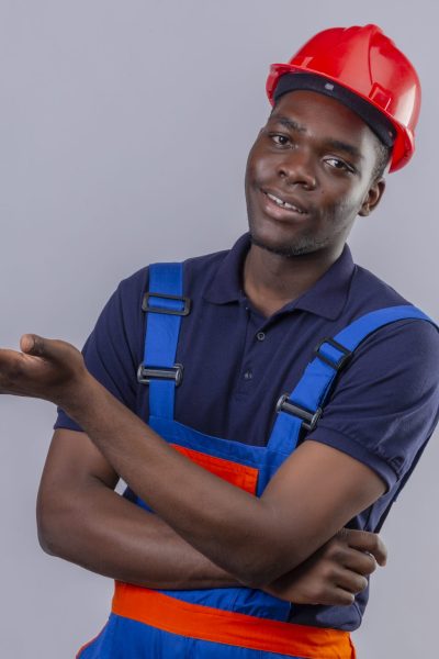 young african american builder man wearing construction uniform and safety helmet pointing with palm of hand looking at camera with smile standing over white background